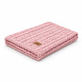 A folded pink chunky knit blanket with a small brown leather brand patch, shown on a solid white background.